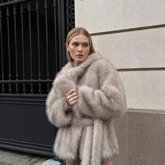 Woman wearing a light gray fur coat standing against a building.