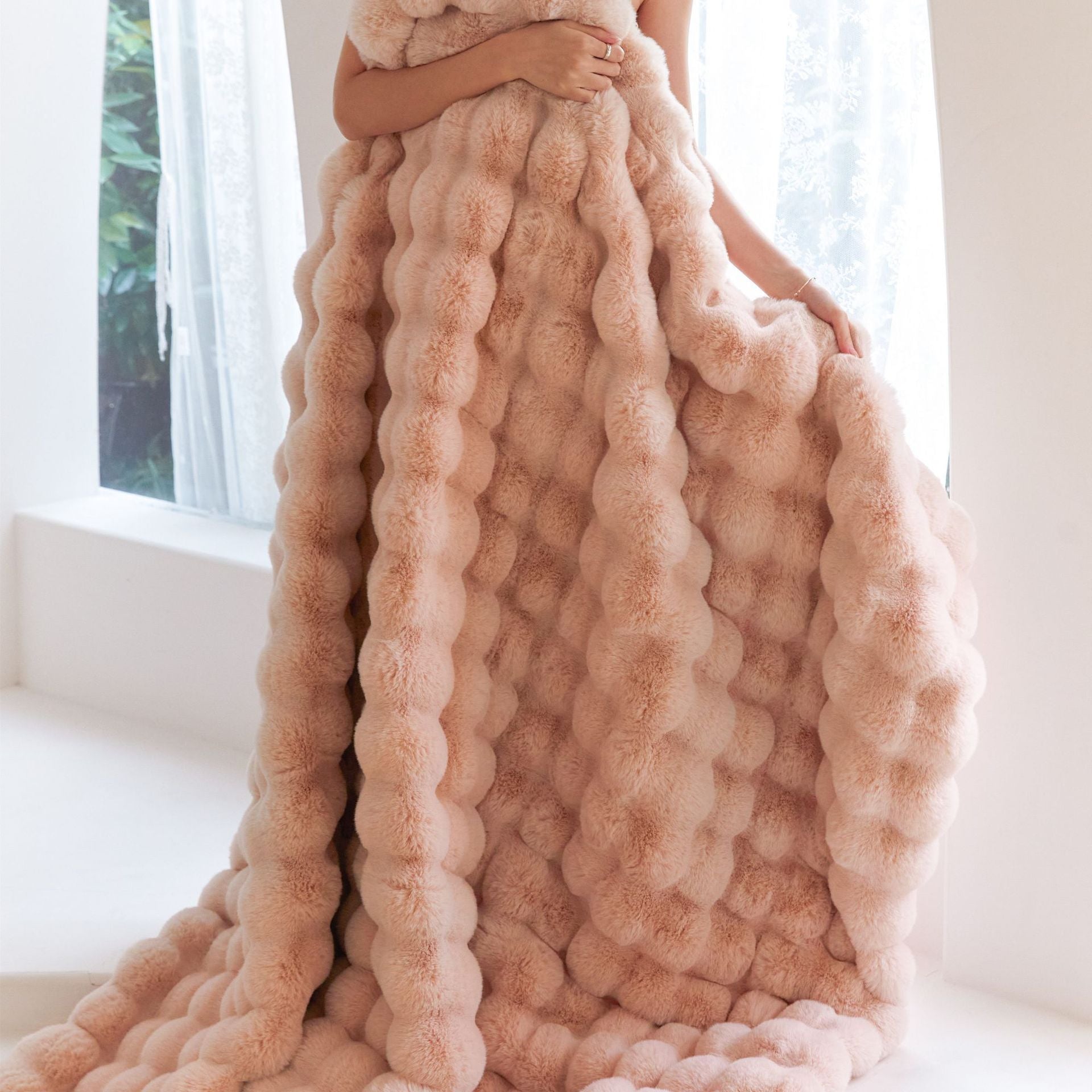 Person wrapped in a large, textured pink blanket in a bright room with white curtains.