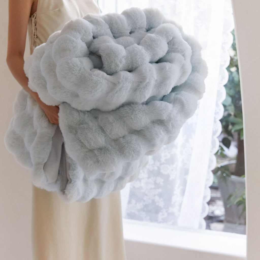 Person holding a large, fluffy gray pillow in front of a window with white curtains.