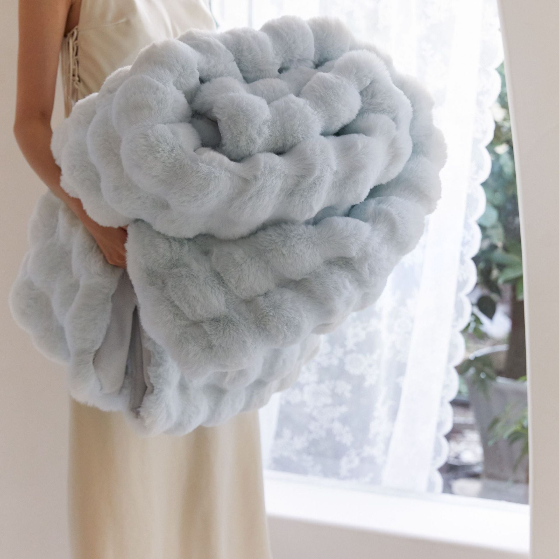 Person holding a large, fluffy gray pillow in front of a window with white curtains.