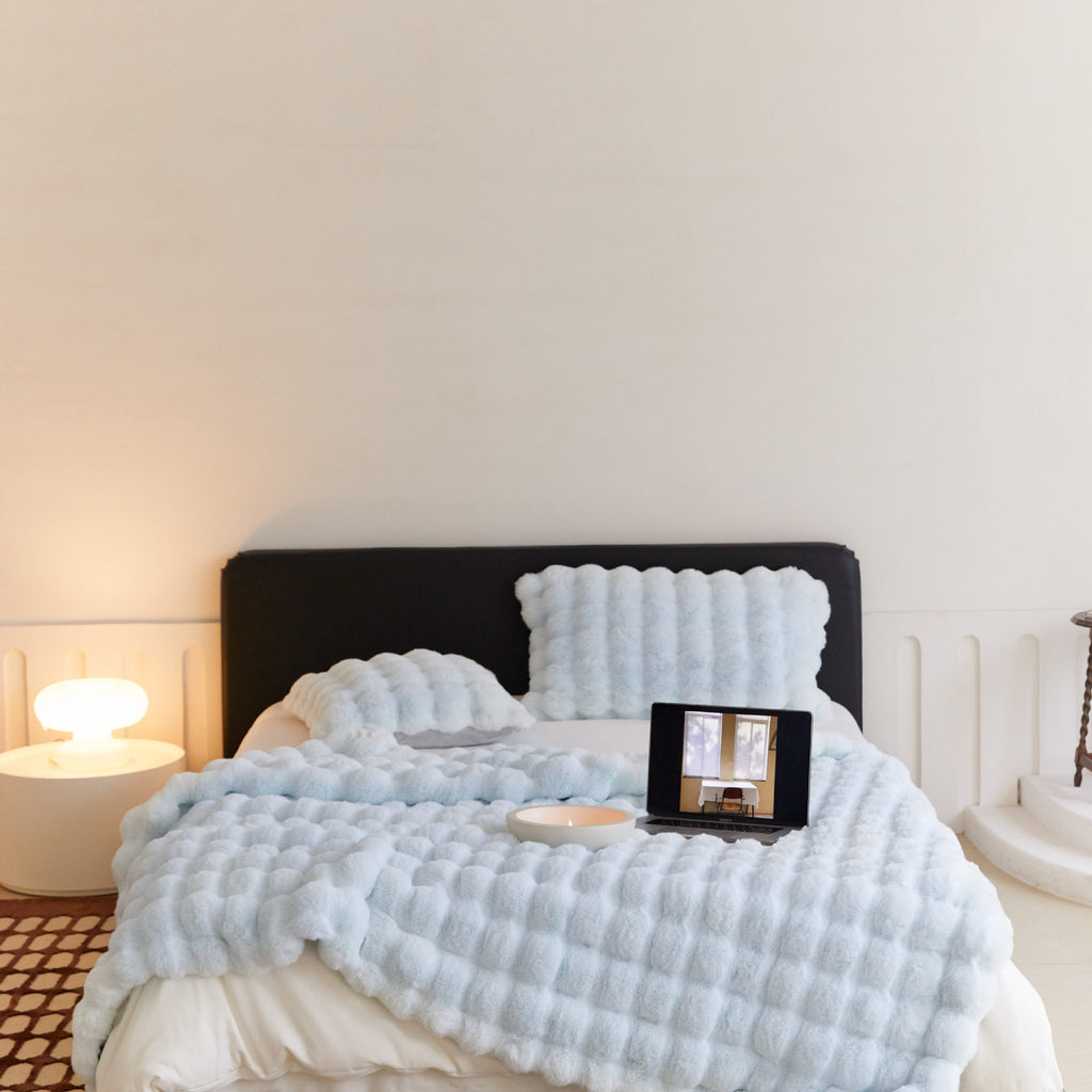 Light blue textured blanket draped over a bed with a lamp and mirror in the background.