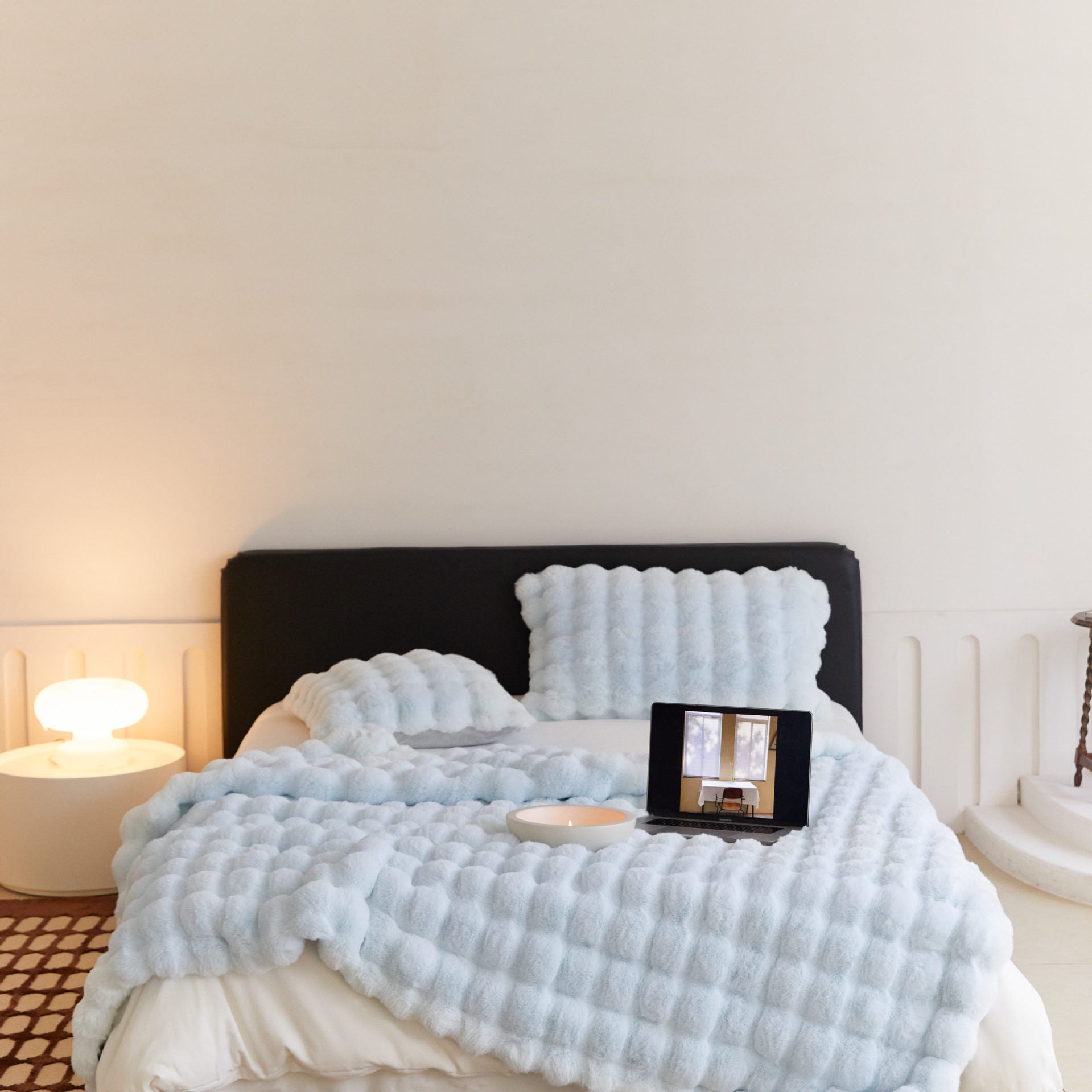 Light blue textured blanket draped over a bed with a lamp and mirror in the background.