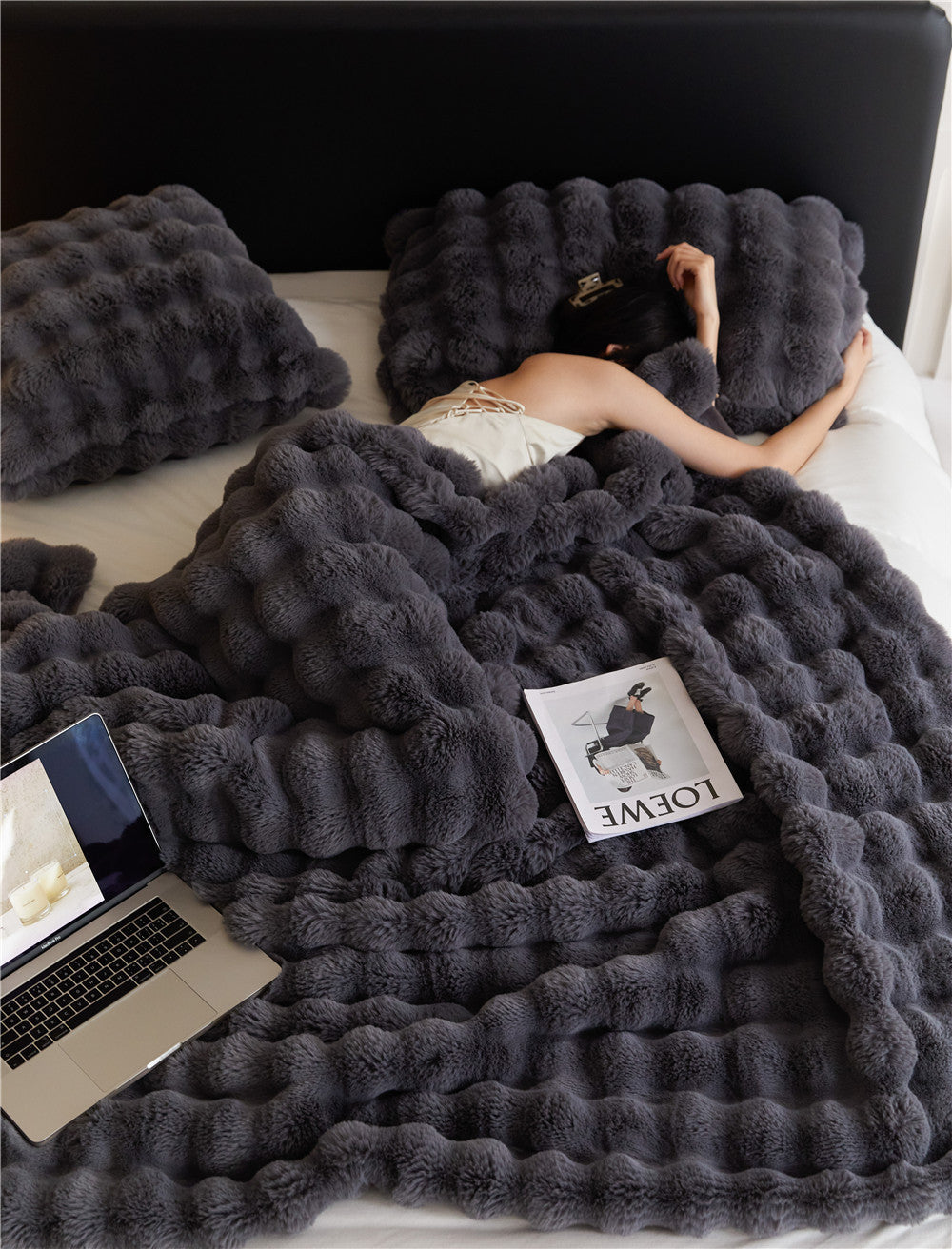 Person lying on a bed under a fluffy gray blanket with a laptop and magazine.