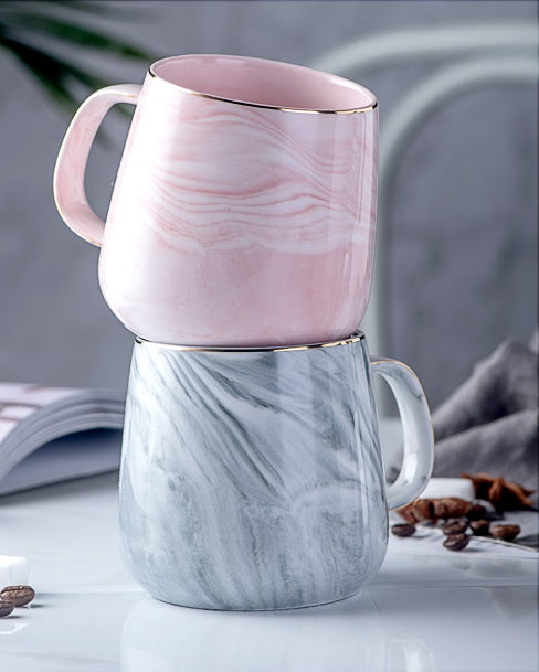 Two marble-patterned mugs, one pink and one gray, on a light surface with coffee beans and a book in the background.