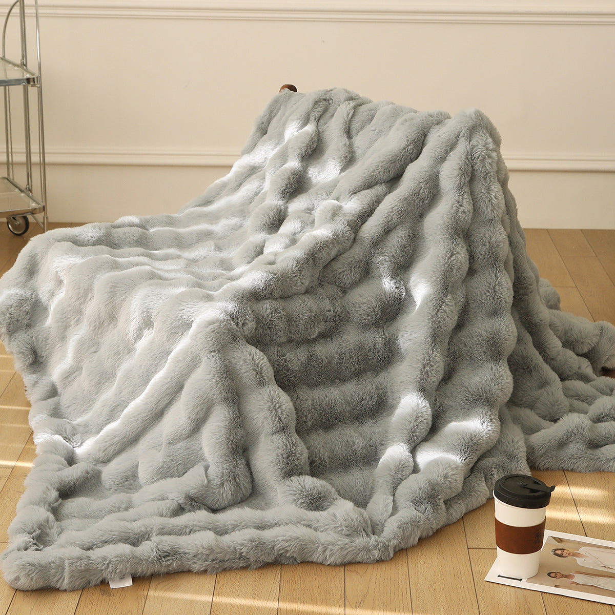 Gray fluffy blanket on a wooden floor with a coffee cup and books in the background.